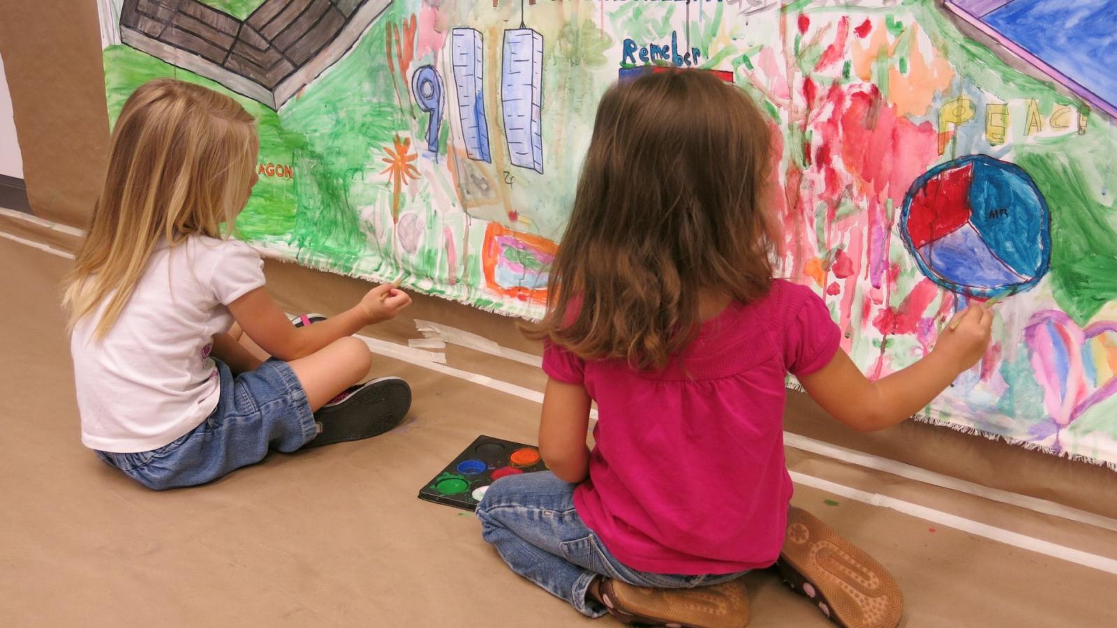 Two small girls, seated, paint a mural on a wall
